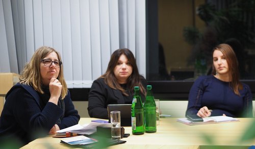 drei Frauen sitzen an einem Tisch mit Unterlagen und hören interessiert zu 