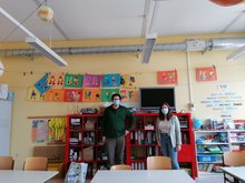 Ein Mann und eine Frau stehen in einem bunt gestalteten Klassensaal einer Grundschule. An der Decke verläuft das Rohr einer Lüftungsanlage.